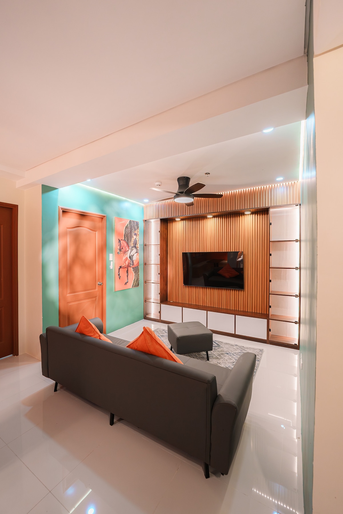 A modern living room features a gray sofa with orange accent pillows positioned in front of a wall-mounted television. A light wood paneling adds texture, and a ceiling fan circulates air in the space. Soft lighting enhances the room's inviting feel.