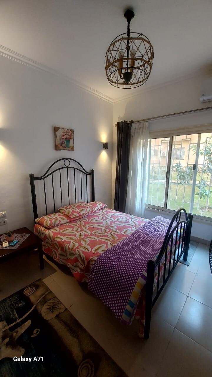 A cozy bedroom features a black metal bed frame with a floral patterned bedspread, accompanied by a colorful polka-dot blanket. Natural light filters through a large window, which offers a view of the garden. A small side table is positioned next to the bed.