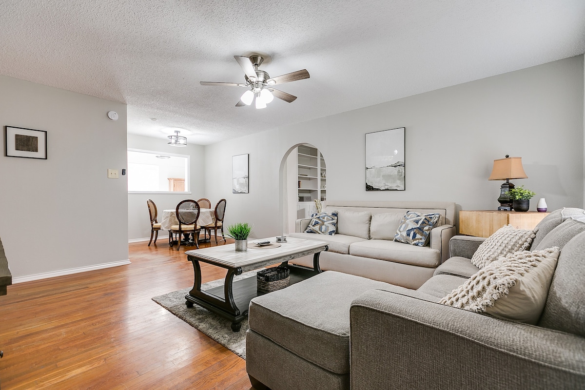 A spacious living area is presented with a comfortable sectional sofa and a coffee table in the center. Light fixtures brighten the room, while a dining area with round chairs is visible in the background. Neutral colors create a calm ambiance, complemented by framed artwork on the walls.