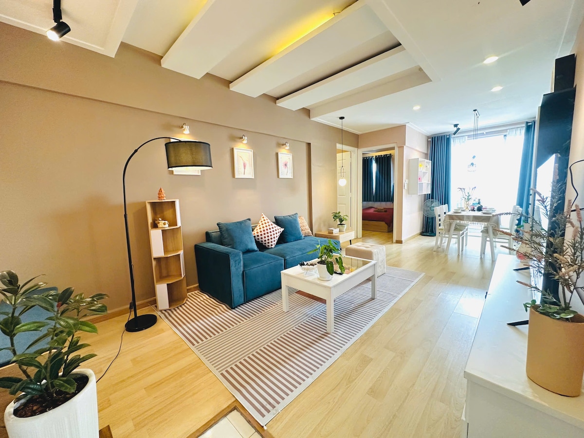 A living area is accented by neutral-toned walls and hardwood flooring. A teal sofa is positioned alongside a white coffee table, with potted plants nearby. Soft lighting illuminates artwork on the walls. A dining space and sleeping areas are visible in the background, with large windows allowing natural light.