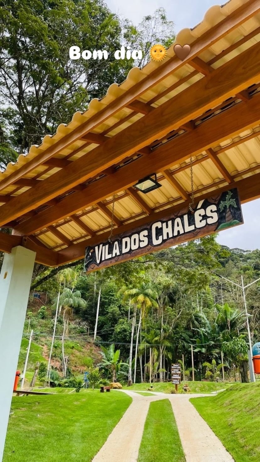 An inviting entrance to Vila dos Chalés is framed by a wooden awning. The sign hangs prominently above a pathway that leads through lush greenery and palm trees, creating a serene atmosphere. A gentle slope of the land can be seen in the background.