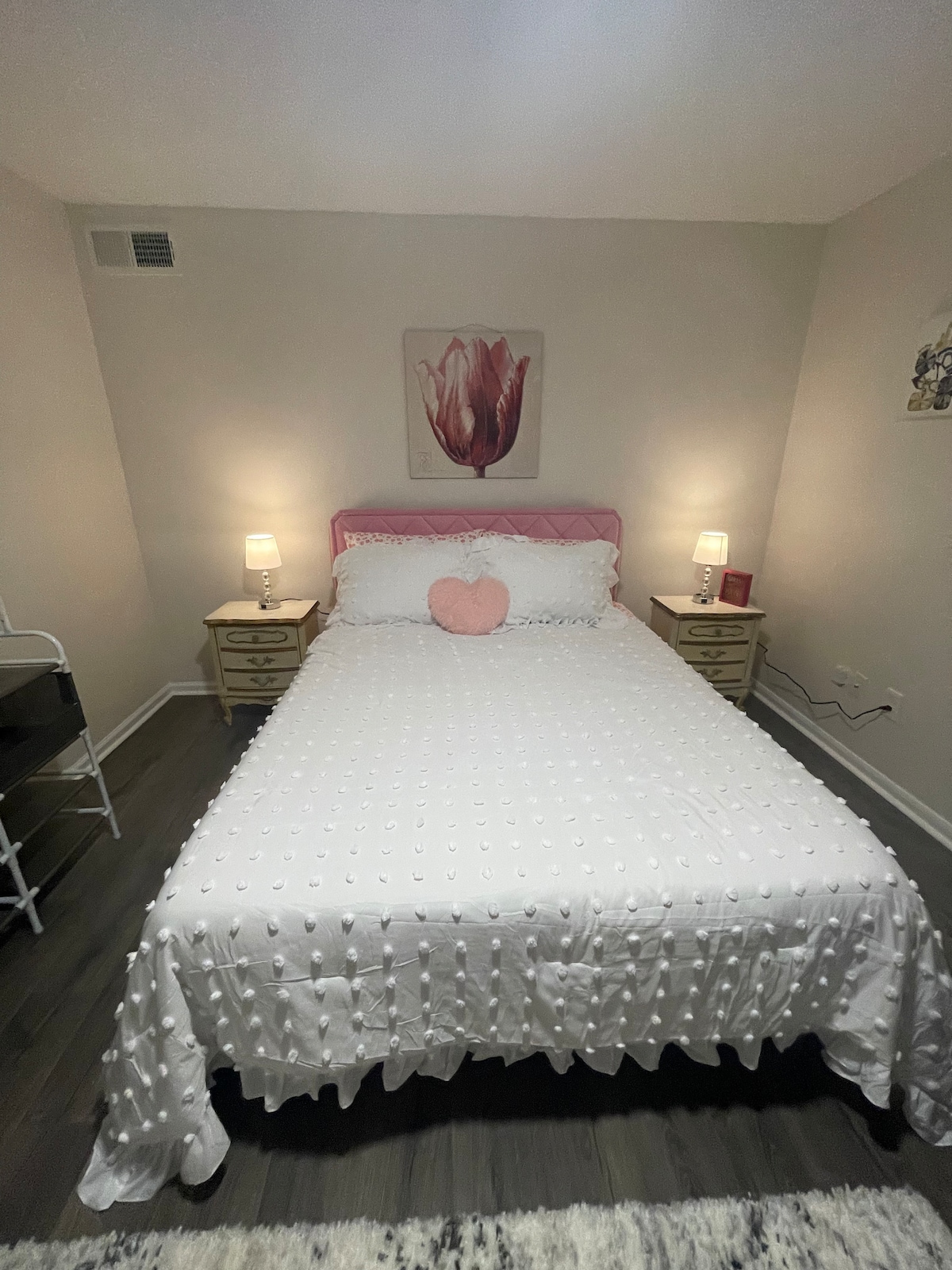 A cozy bedroom features a spacious bed adorned with a textured white coverlet and a decorative heart-shaped pillow. Soft bedside lamps illuminate the area, while a floral art piece is displayed above the headboard. Light wood nightstands are positioned on either side of the bed.