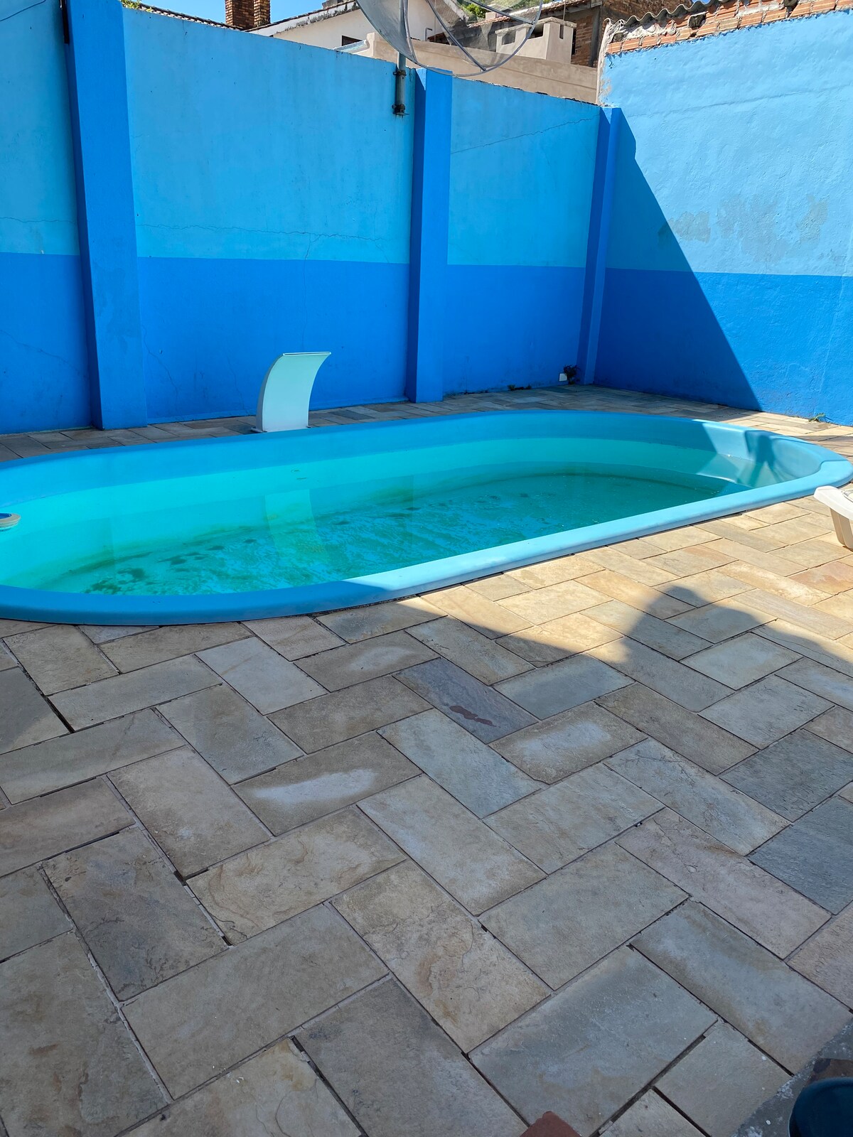 A rectangular pool is set within a blue-walled area, featuring a smooth, curved edge. The water appears calm, surrounded by a light-colored stone patio that adds texture to the space. Sunlight casts a soft shadow on the ground.