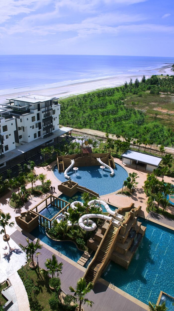 An aerial view reveals a large swimming pool area featuring a water slide. Sun loungers and palm trees surround the pools, providing a tropical feel. The beach and sea are visible in the background, with greenery lining the coastal area and a modern building on the left.