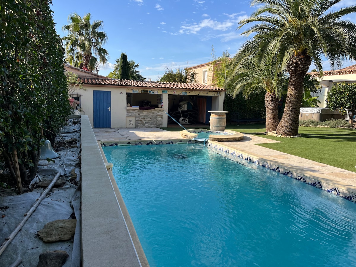 A turquoise pool is featured in a private garden setting, surrounded by palm trees and lush greenery. An outdoor kitchen area is visible, along with a stone wall and a barrel. Clear blue skies reflect a serene atmosphere over the inviting space.