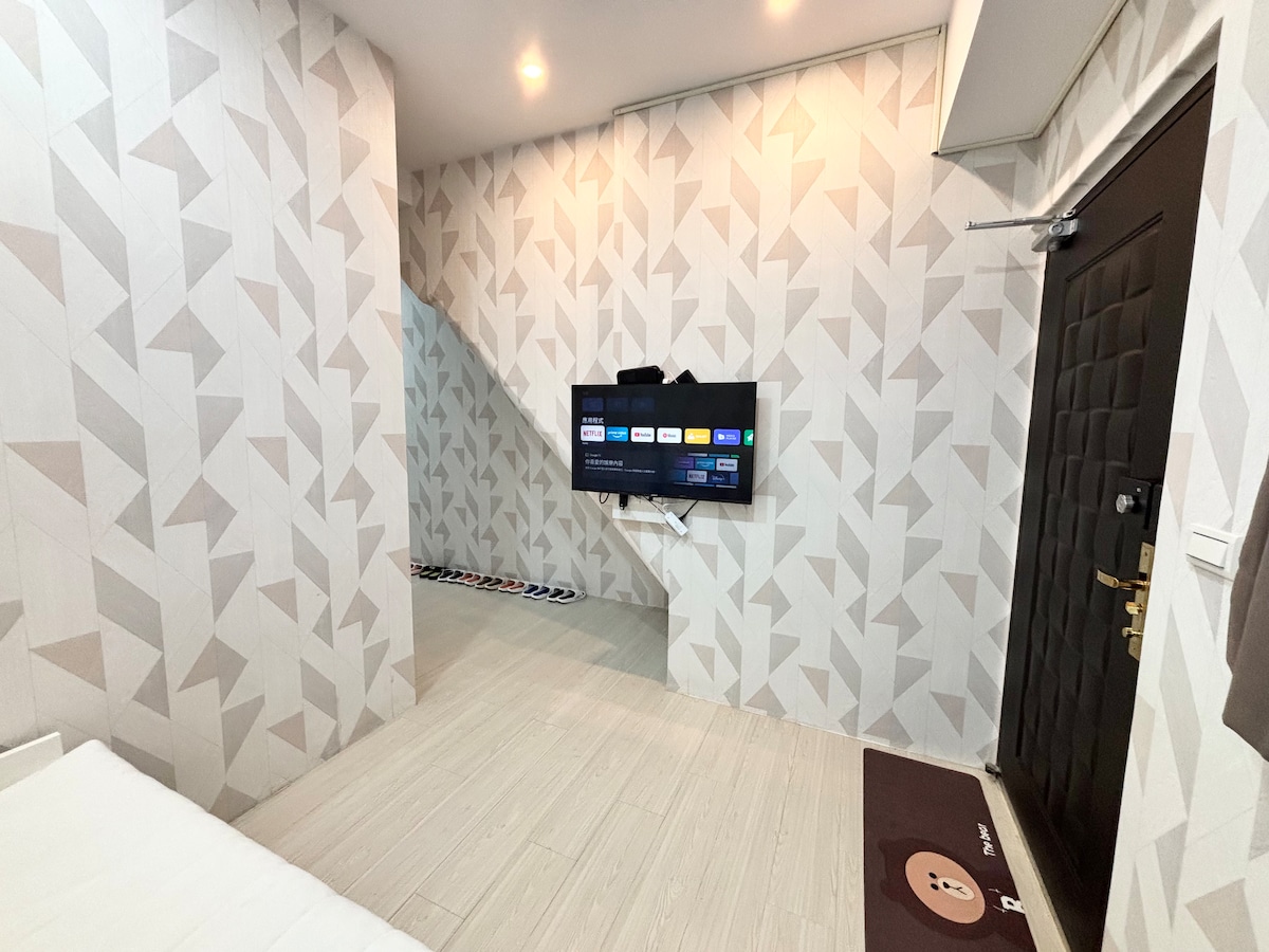 A well-lit room features geometric patterned walls and light-colored flooring. A flat-screen television is mounted on the wall, opposite a sleeping area that includes a spacious bed. A door on the right leads to an entrance, while a welcome mat is positioned at the front.
