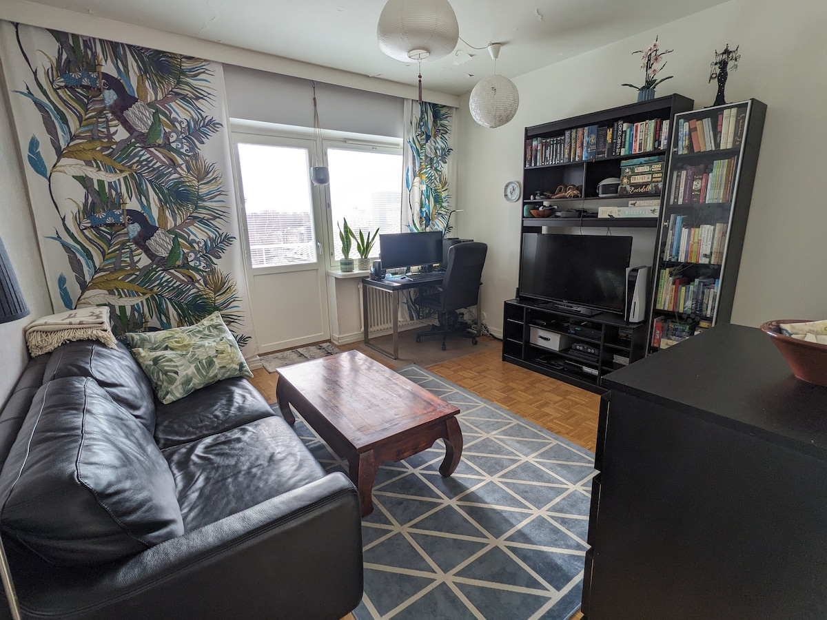 A living room is depicted, featuring a dark leather sofa paired with a wooden coffee table. A bookshelf filled with various books and decorative items occupies one side. Large windows allow natural light to brighten the space, accented by vibrant foliage-patterned curtains. A workspace is positioned in the corner.