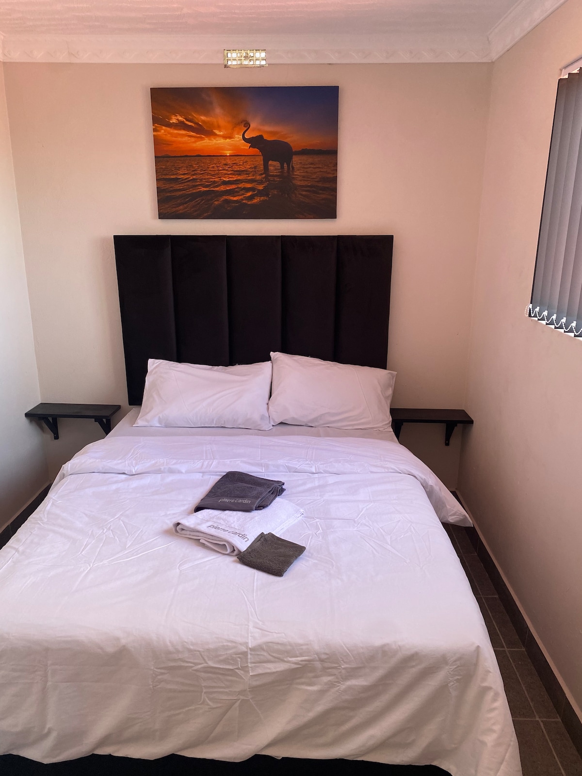 A compact sleeping area features a double bed with white bed linen and towels neatly arranged. Two side shelves are positioned beside the bed. A wall art piece showcasing an elephant against a sunset adds a serene touch, while window blinds allow for adjustable light.