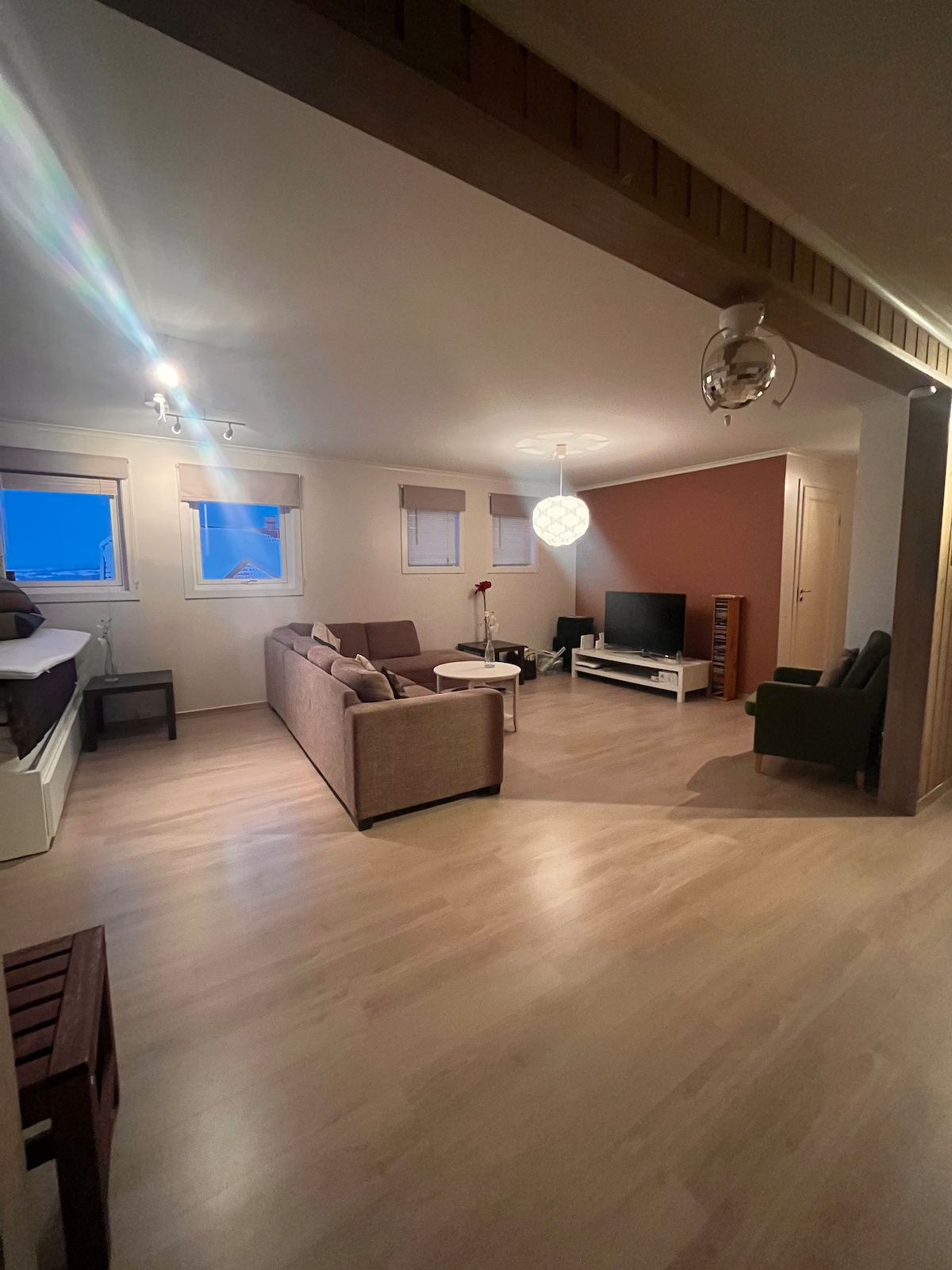 The open living area features a comfortable seating arrangement with a sofa and armchairs. A television is positioned against a white media unit. Natural light enters through multiple windows with blinds, illuminating the light wood flooring and softly colored walls.