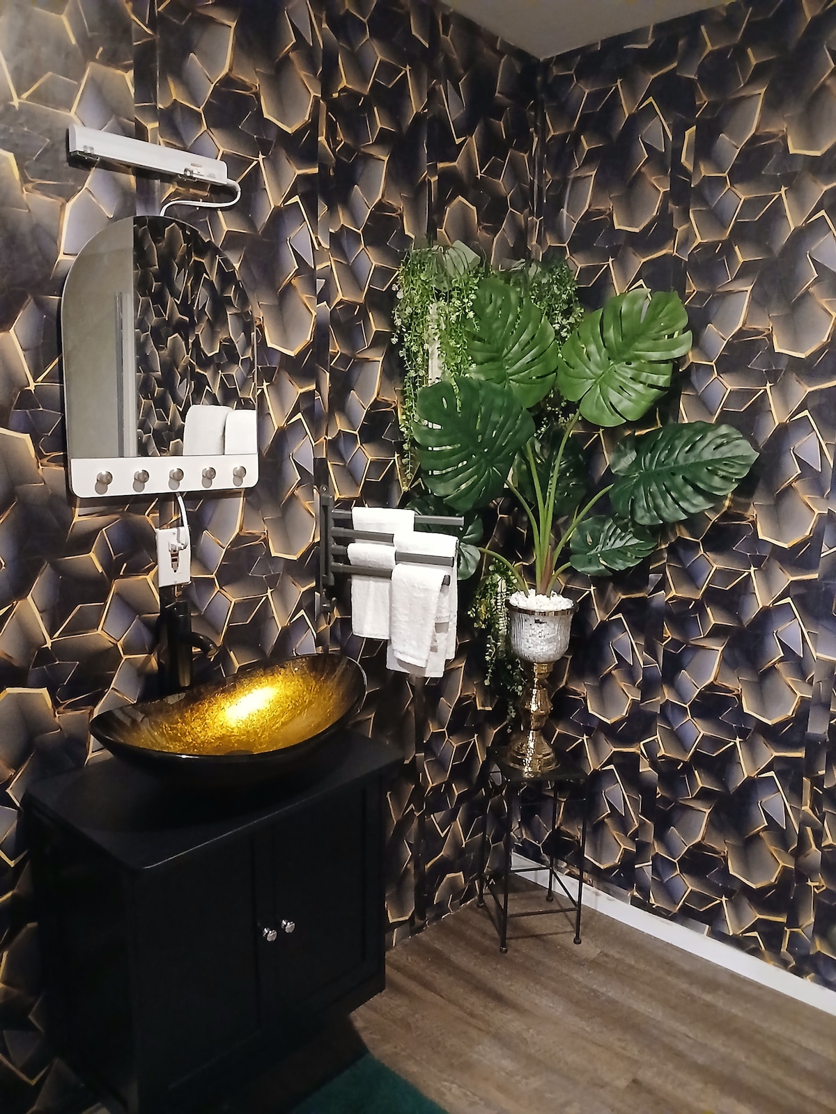 The bathroom features an elegant black and gold geometric wall design, complemented by a stylish black vanity with a gold sink. Soft towels are neatly arranged on a wall rack, and a decorative plant adds a touch of greenery to the space.