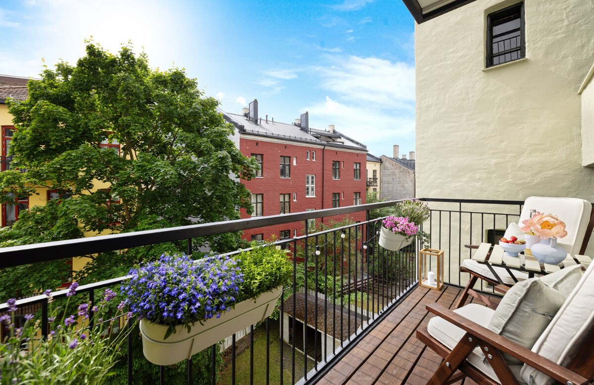 A balcony is seen adorned with colorful flower pots and two reclining chairs. Sunlight bathes the outdoor space, highlighting views of neighboring buildings and lush greenery. The wooden deck provides a cozy area for relaxation amid a peaceful urban setting.