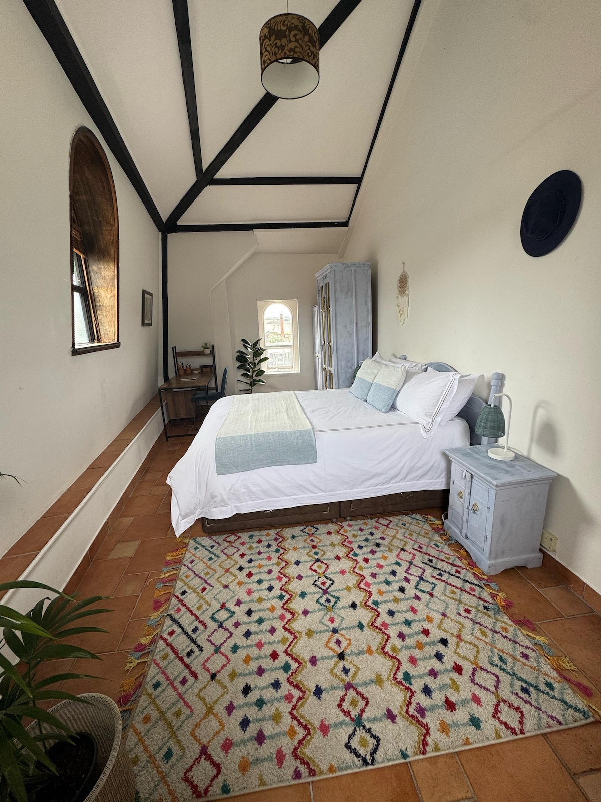 A rectangular bedroom features a spacious bed with light bedding, flanked by a bedside table and a lamp. A colorful area rug adds warmth to the terracotta tile floor. A window provides natural light, and a chair is positioned near the entrance.