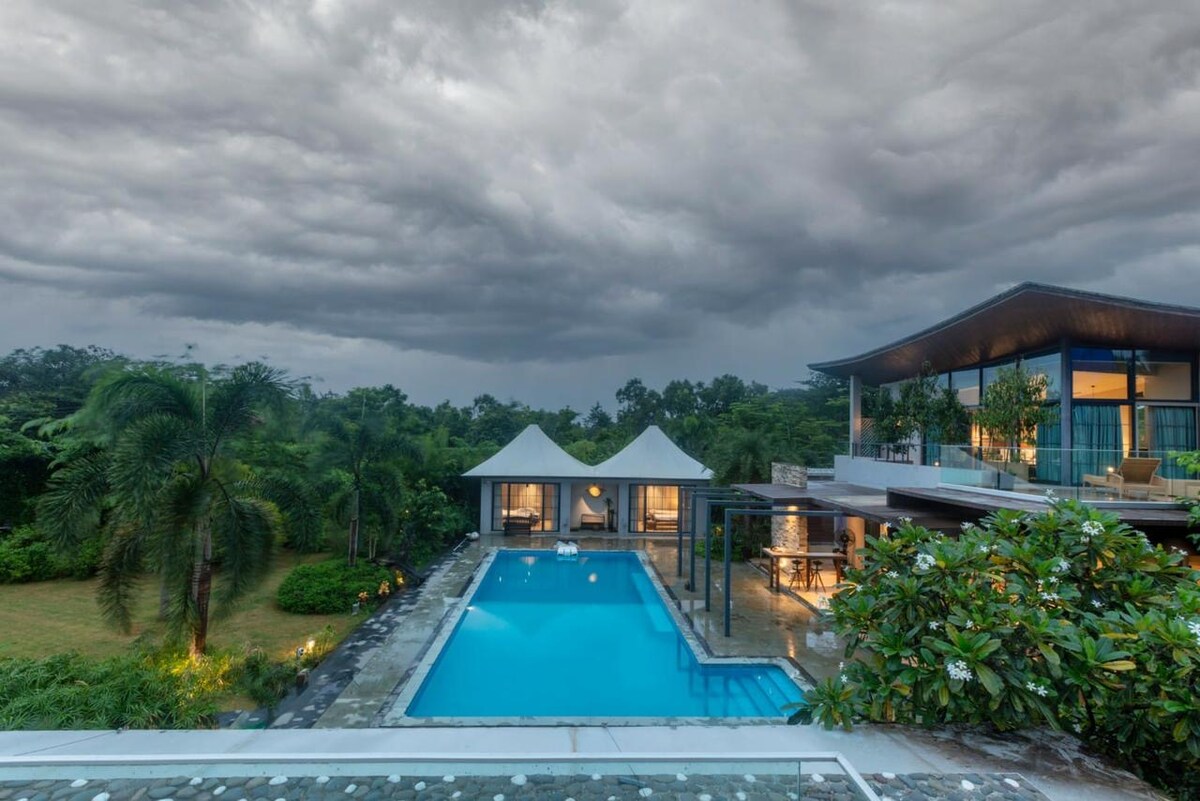 A spacious outdoor area features a large swimming pool surrounded by lush greenery. Two shaded cabanas are positioned near the pool, while a modern villa stands to the right, showcasing expansive windows and a covered dining space. The sky is overcast, hinting at an impending rain.