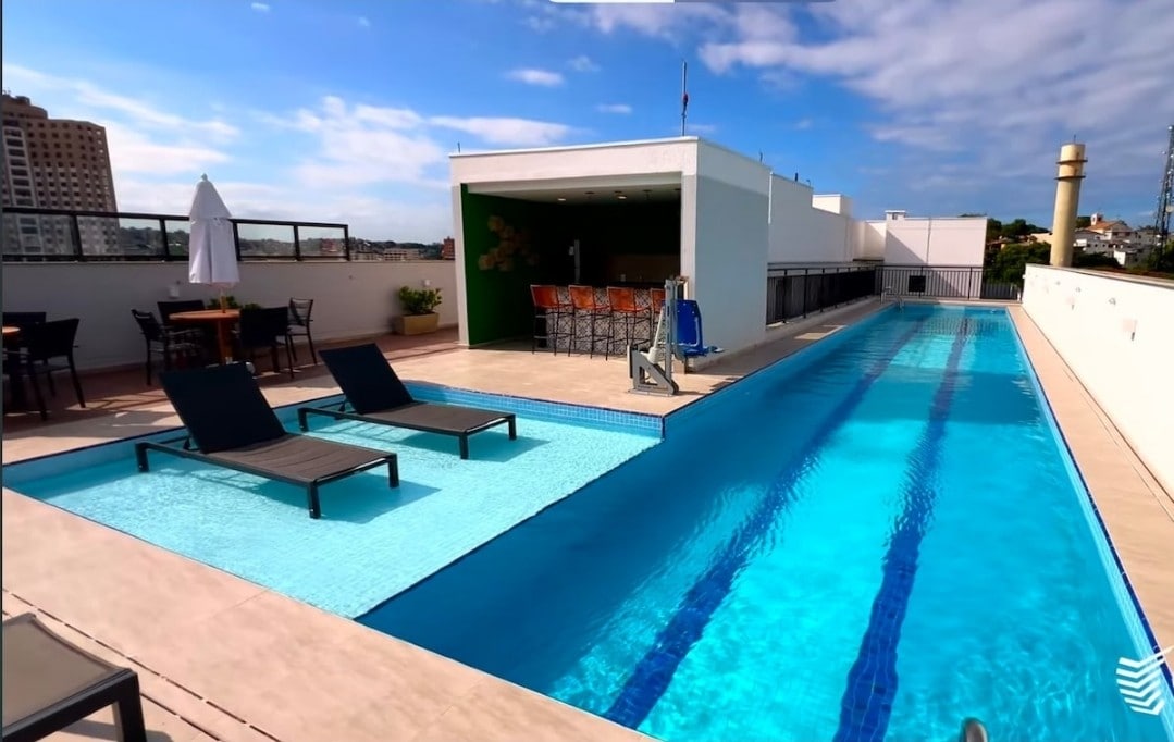 A rooftop area features a long swimming pool bordered by lounge chairs. A shaded seating area with tables and chairs is visible in the background. The space is well-lit with natural sunlight and offers expansive views of the surrounding city.