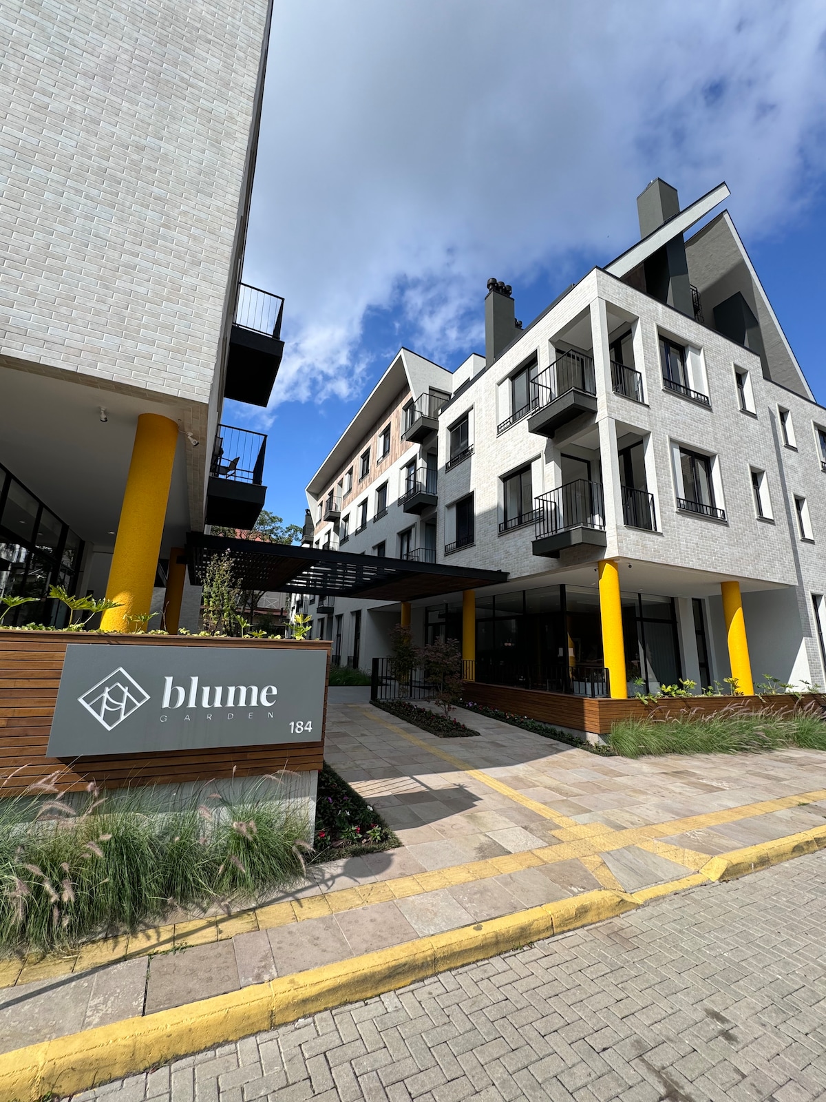 The exterior of the condominium is presented, showcasing a modern design with multiple levels and balconies. A welcoming entrance with a sign reading 'blume' is framed by decorative landscaping and cheerful yellow columns. The clear blue sky adds a vibrant touch to the overall aesthetic.