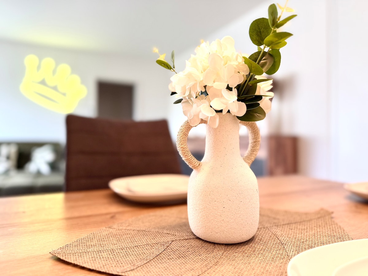 A decorative vase with a textured surface holds fresh white flowers, placed on a woven placemat. The dining table, featuring natural wood tones, is set with simple plates, contributing to a clean and inviting atmosphere in the dining area.