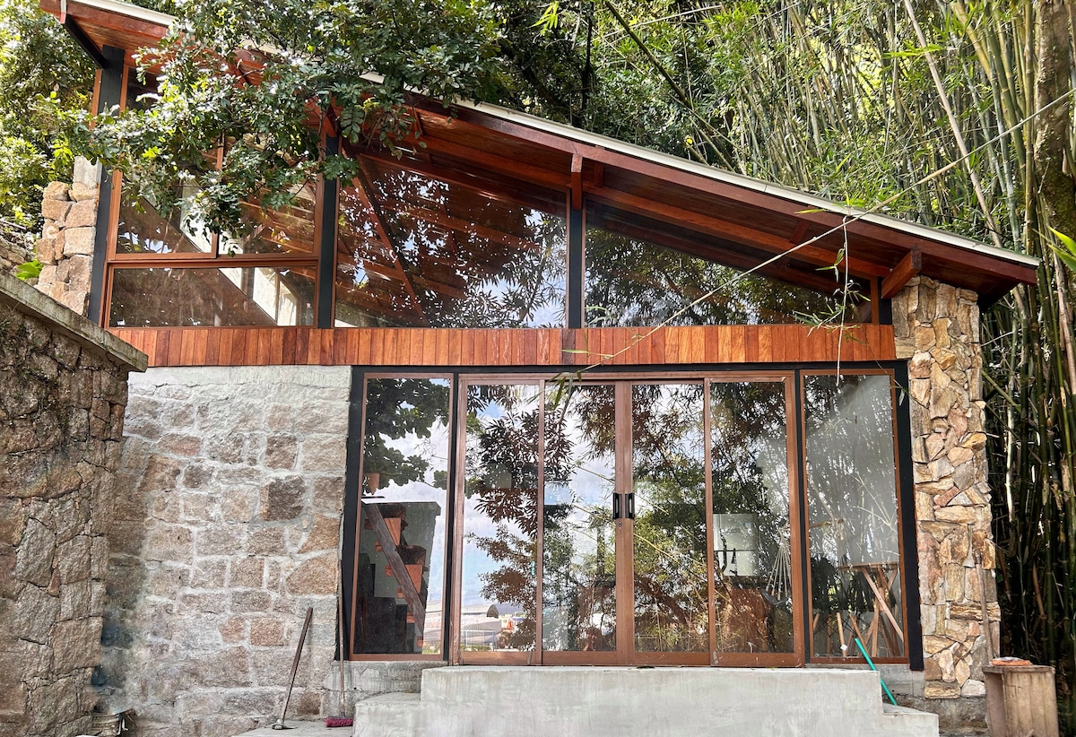 A charming chalet features a blend of stone, wood, and glass in its construction. Large glass doors allow natural light to fill the space, while bamboo and trees surround the area, creating a serene connection with nature.