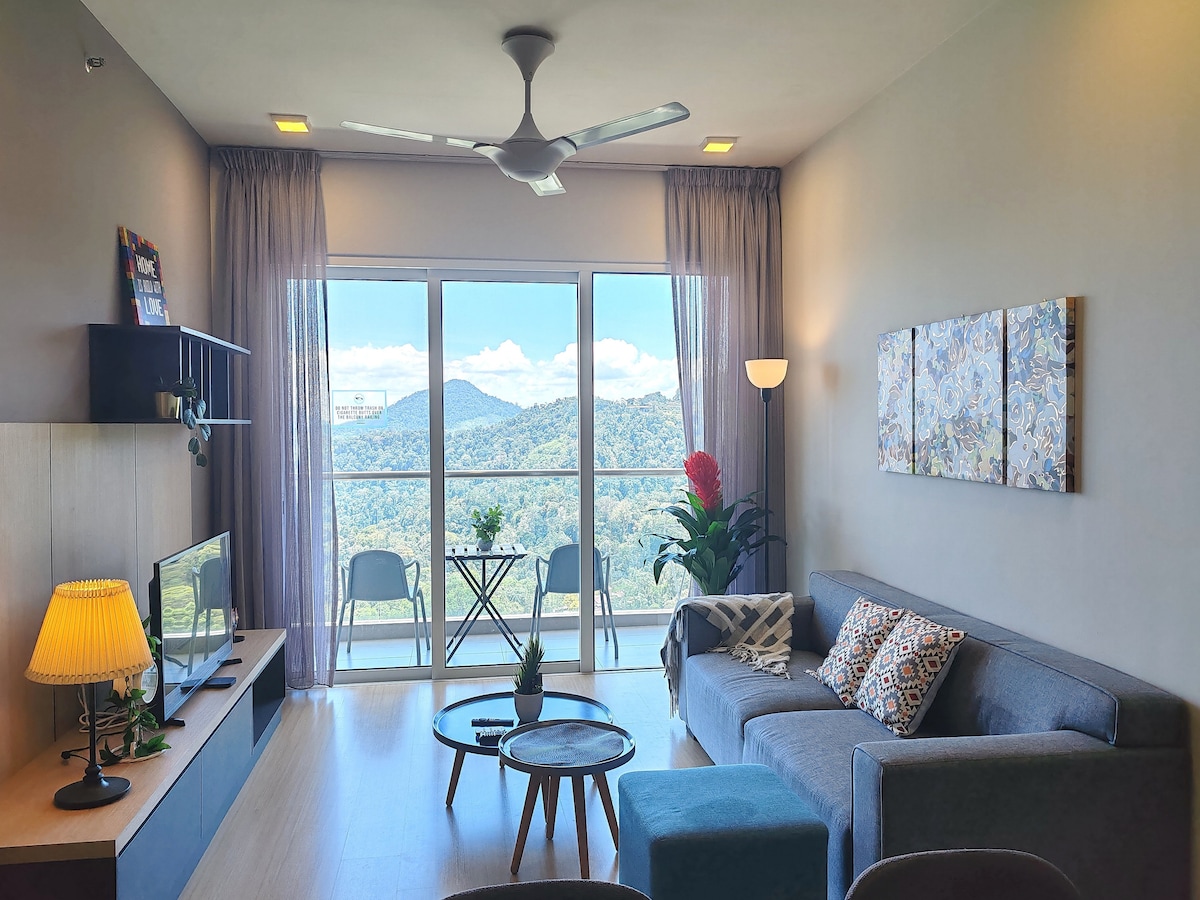 A bright living area features a comfortable gray sofa and a round coffee table. Two soft armchairs are arranged beside a television unit. Large glass doors open to a scenic balcony, with mountains visible in the background. Soft lighting is provided by a standing lamp.