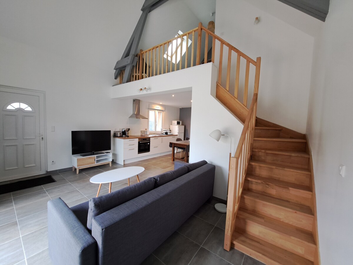 A spacious living area features a comfortable sofa facing a white coffee table. A staircase with wooden railings leads to the duplex sleeping area. The kitchen is visible in the background, equipped with modern appliances and ample counter space. Natural light enters through the window above.