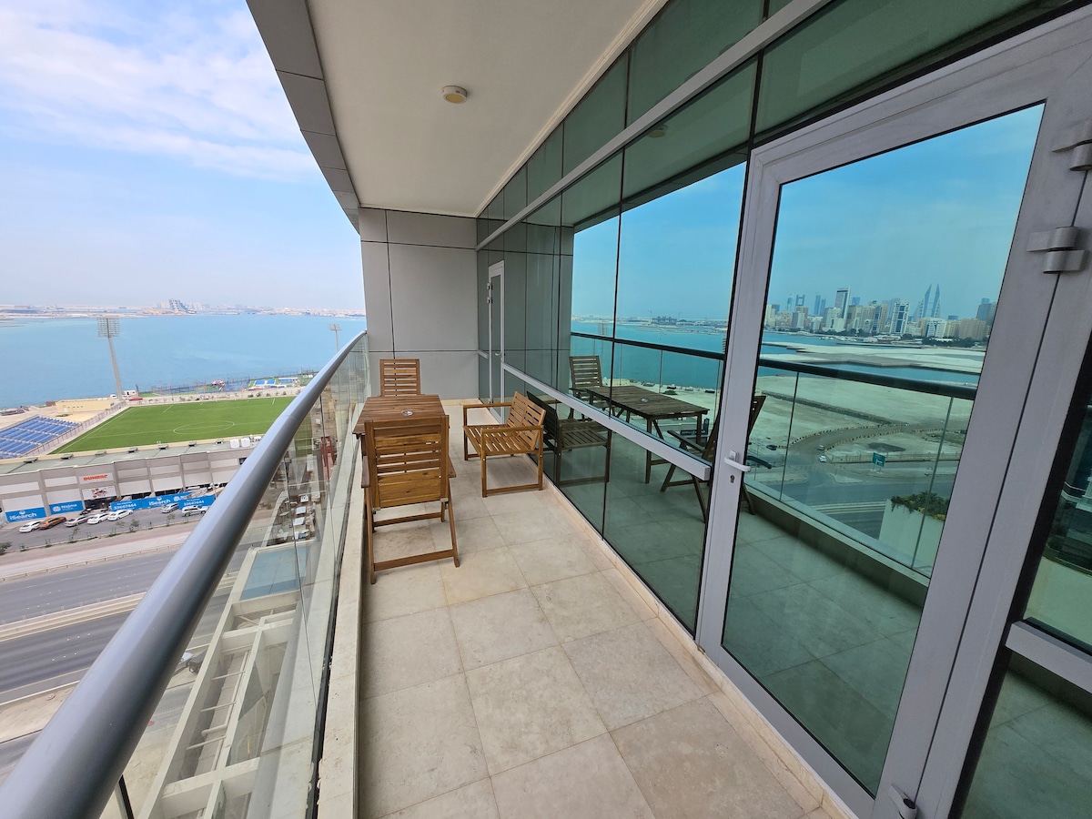 A spacious balcony offers stunning views of the water and city skyline. Two wooden chairs and a matching table are arranged on the tiled surface, reflecting the scenery through the large glass doors. The urban landscape is visible in the distance.