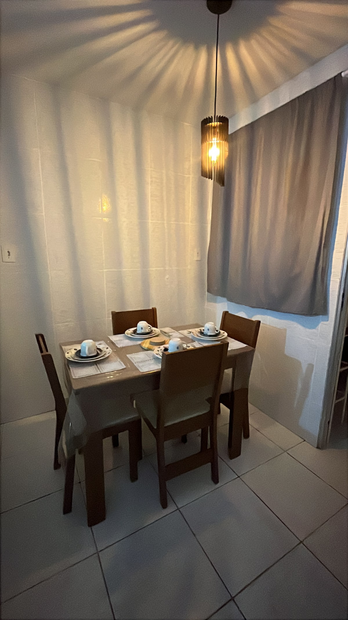 A dining area is presented with a square table set for four, accompanied by wooden chairs. Soft lighting is provided by a pendant fixture above. Light-grey curtains partially cover a window, allowing gentle illumination to fill the space.