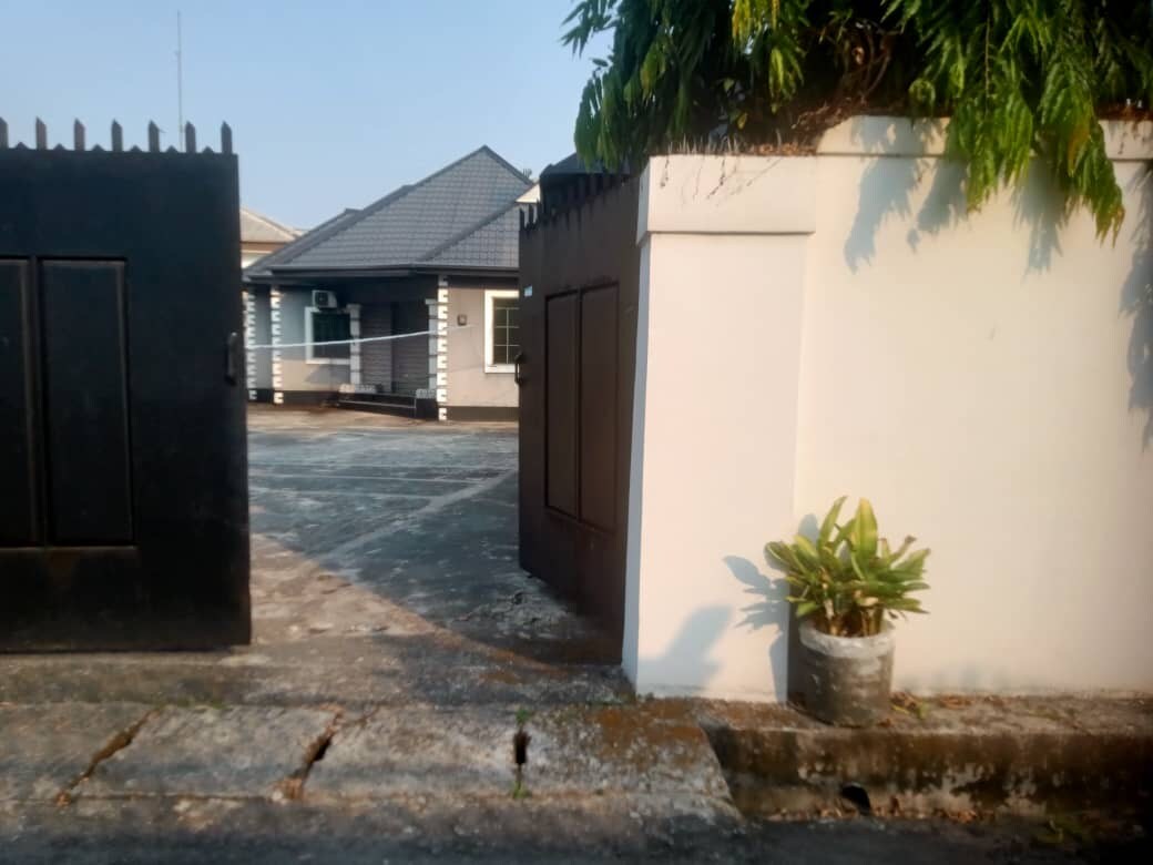 The entrance features a pair of contrasting gates, one black and one white, leading into a paved area. A small potted plant sits beside the gate, with a glimpse of a well-maintained building visible in the background. The sky is clear, enhancing the overall brightness.