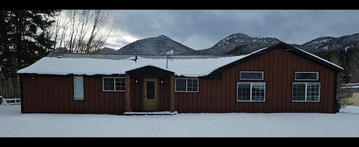 A spacious brown house is set against a backdrop of snow-covered mountains, with a light dusting of snow on the ground. Large windows reflect the cloudy sky, and a welcoming front door is centered on the facade, inviting guests into the cozy retreat.