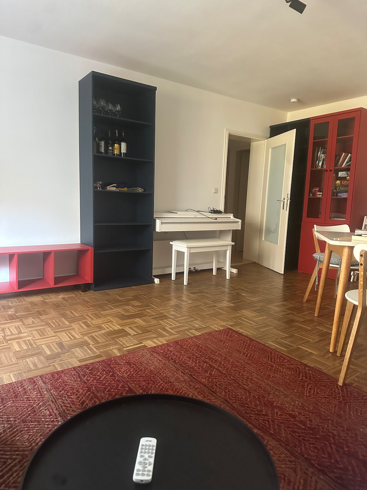 A bright interior is presented, featuring a wooden floor and colorful furniture. A tall black shelving unit holds glasses and bottles. A white desk sits in front of a doorway, while a red cabinet is positioned beside it. A round black table is visible in the foreground.