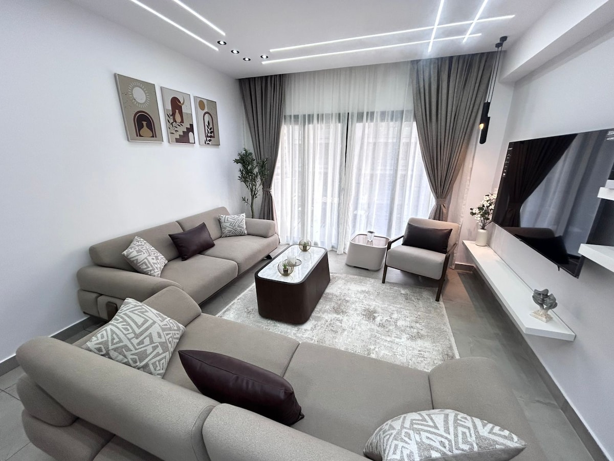 A modern living area is arranged with a light-colored sectional sofa featuring dark accent pillows. A sleek coffee table is positioned at the center, complemented by a soft area rug. Large windows with sheer curtains allow natural light to fill the room, enhancing the open feel.