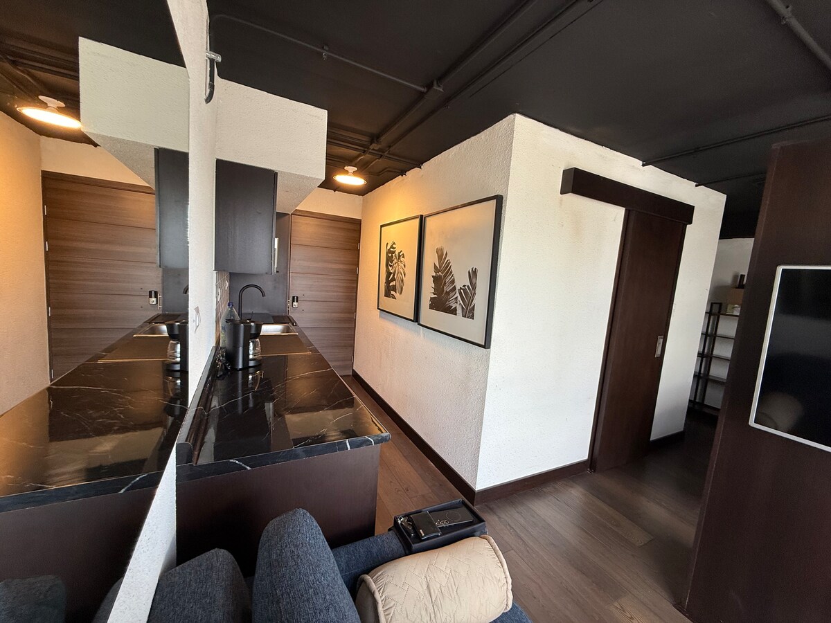An open-concept space features modern kitchen fixtures and dark wood cabinetry. Across the room, framed artwork adorns the walls. A sectional couch invites relaxation, while a dark television console is positioned within view, complementing the sleek design of the loft.