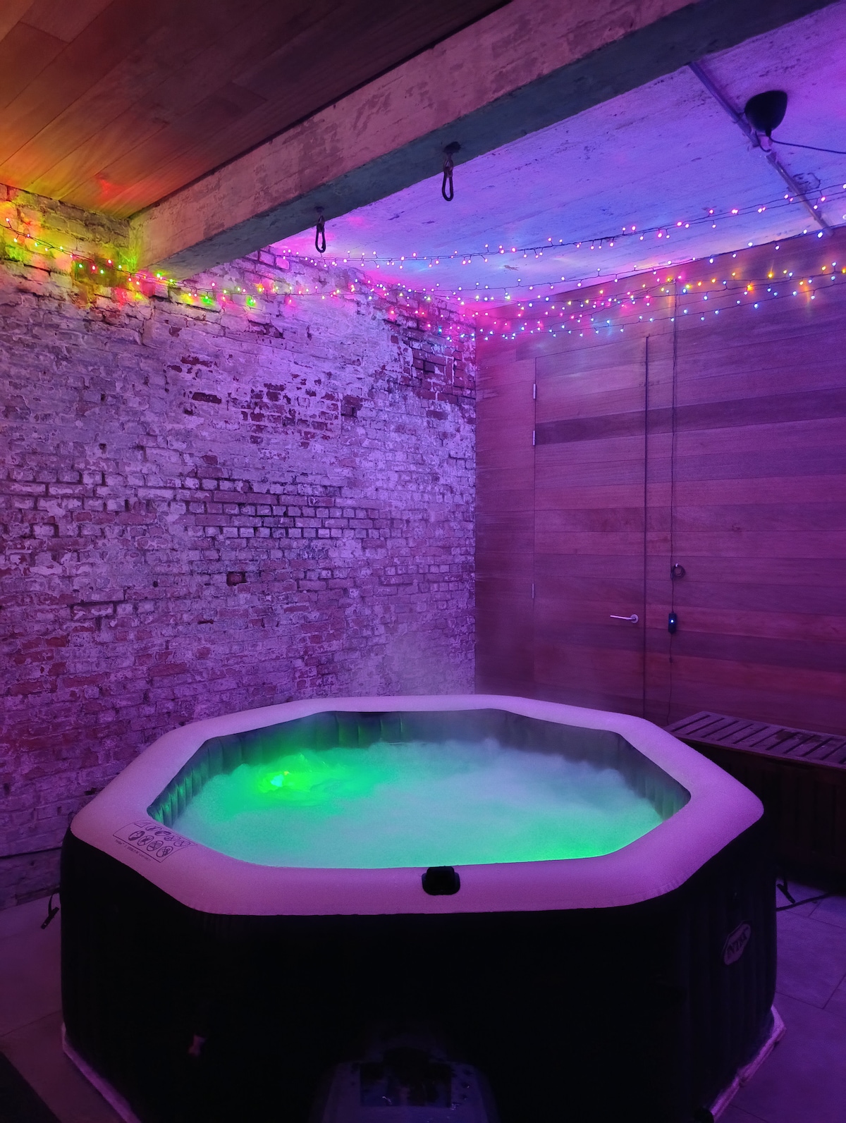 An overed hot tub is presented, surrounded by a rustic brick wall. Colorful lights adorn the ceiling, creating an atmospheric glow. Steam rises gently from the water, which features a soft green hue, inviting relaxation in a private setting.