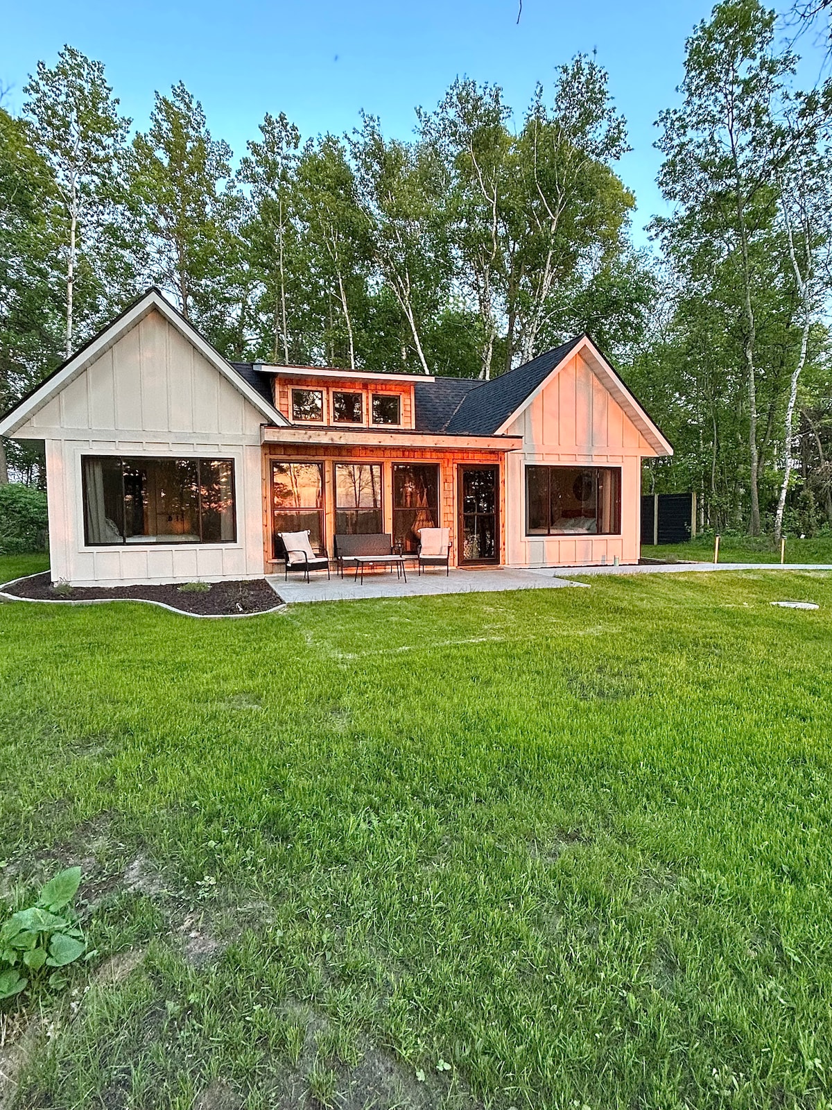 The cabin features a modern exterior with light-colored siding and a black roof. Large windows allow for abundant natural light, and a covered porch is furnished with two chairs. A well-maintained green lawn extends in front, with trees providing a natural backdrop.