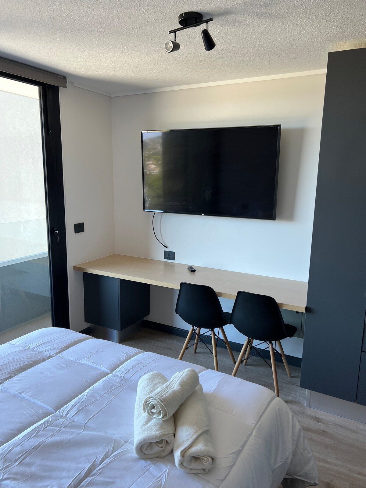 A cozy sleeping area features a spacious bed with rolled towels at the foot. A wall-mounted television is visible above a minimalist desk, accompanied by two chairs. Natural light filters in through a large window, enhancing the serene atmosphere of the room.