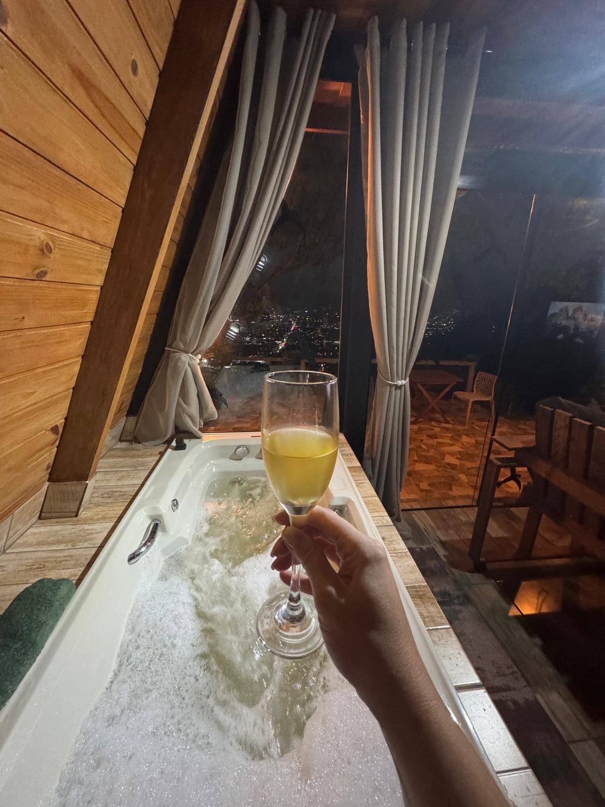 A cozy indoor space is featured, with a bubbling jacuzzi in the foreground. Soft lighting reflects off the water. A hand holds a glass of beverage, which adds a touch of relaxation. Beyond, a view of the city can be seen through large windows draped with light curtains.