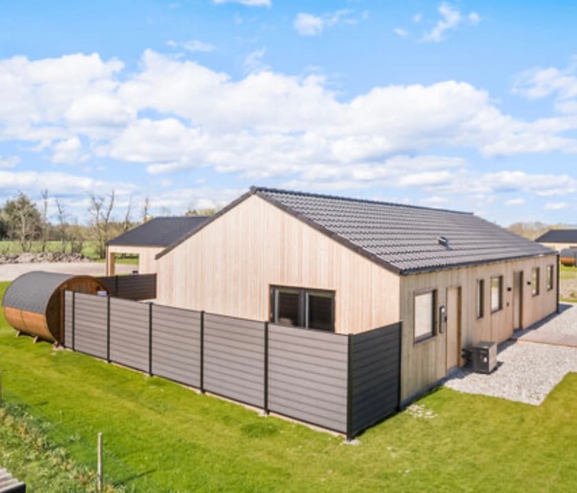 The exterior of a wooden house is viewed from a distance, showcasing a modern design with a sloped roof. A wooden sauna is positioned to the left, while a fenced yard with green grass extends around the property. Blue skies and scattered clouds provide a serene backdrop.