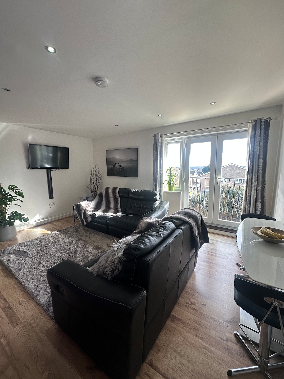 A comfortable living area is presented with a black leather sofa and a matching armchair facing a wall-mounted television. A small dining table with chairs is positioned nearby, and large glass doors lead to a balcony, allowing natural light to fill the space.