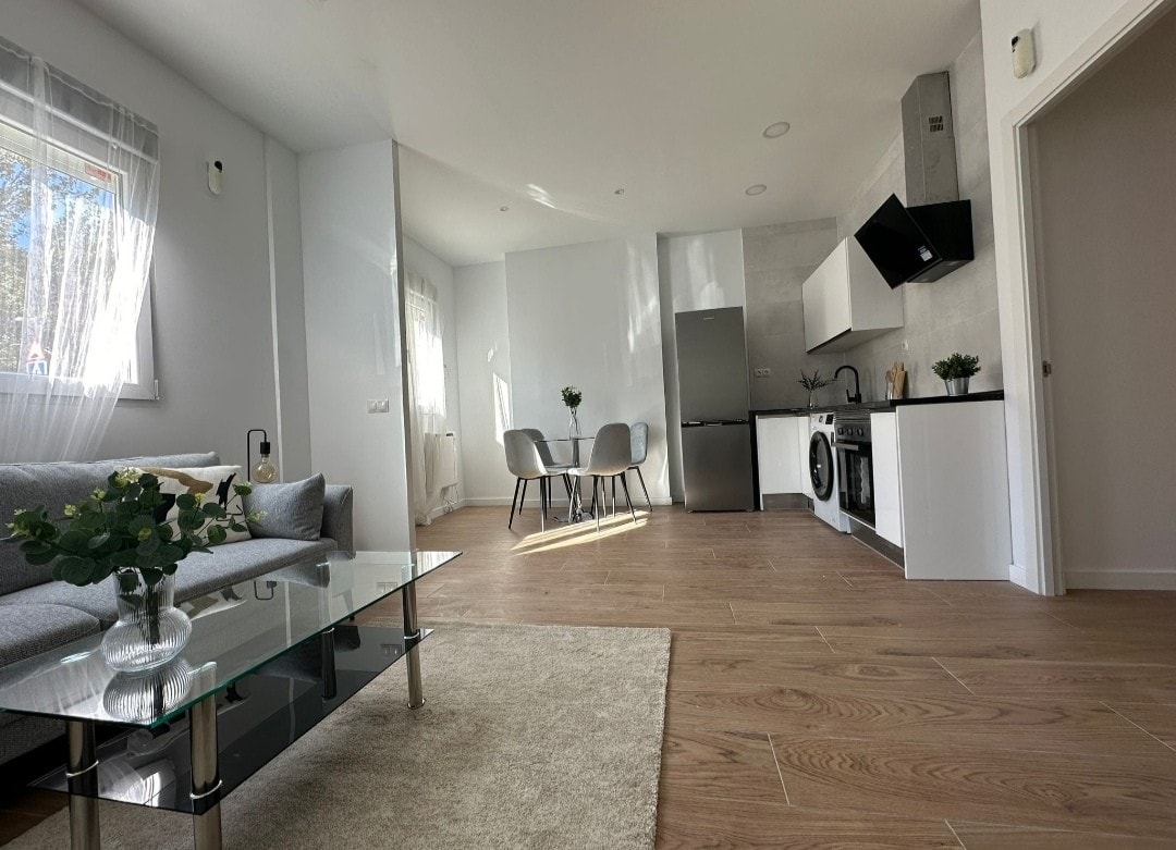 A modern living area features a comfortable gray sofa positioned across from a glass coffee table. Natural light brightens the space through a window, highlighting the open-plan kitchen that includes sleek appliances. A dining table for two complements the inviting atmosphere.