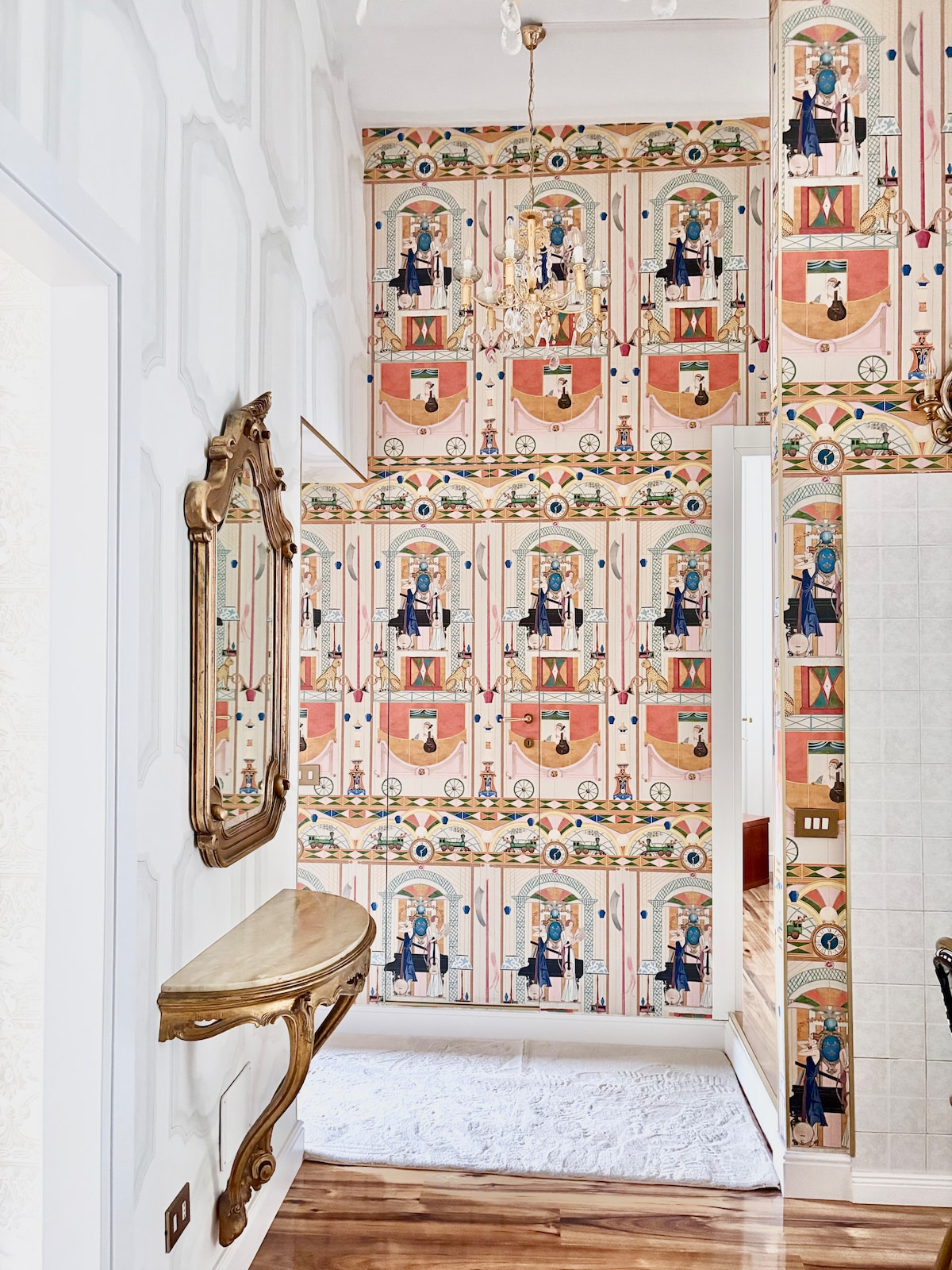 A modern bathroom showcases vibrant, patterned wallpaper featuring artistic figures in bright colors. A small golden console table adds elegance, accompanied by an ornate mirror. Soft lighting enhances the inviting atmosphere, while the wooden floor complements the decorative elements for a stylish and functional space.