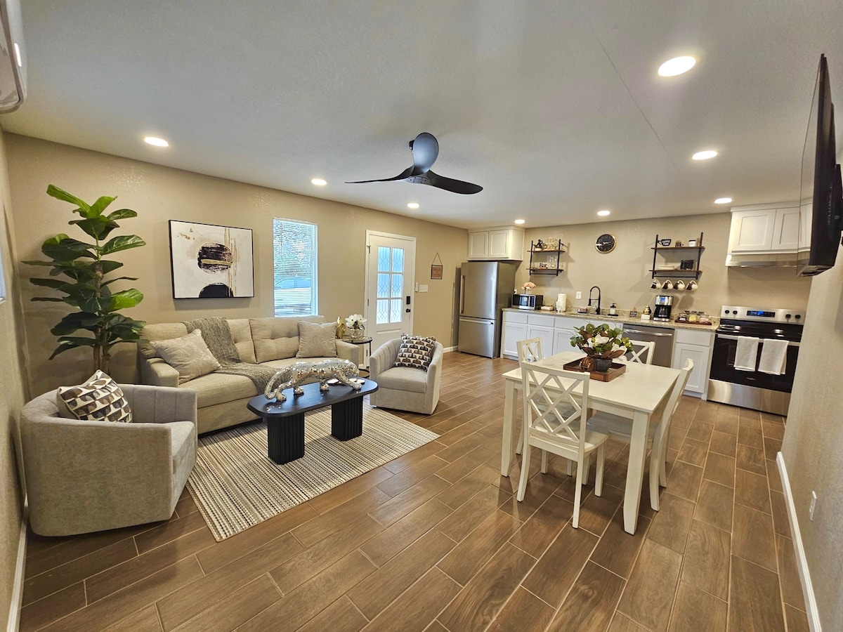 A modern living area features a comfortable gray sofa, accented by decorative pillows, and two black coffee tables. A dining table with four white chairs is positioned nearby. A fully equipped kitchen is visible in the background, alongside soft overhead lighting and natural light from the windows.