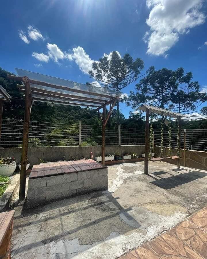 An outdoor area features wooden structures with shaded seating. The space is surrounded by greenery, with a concrete surface and decorative plants in pots. Above, a clear sky and sunlight create a bright atmosphere.
