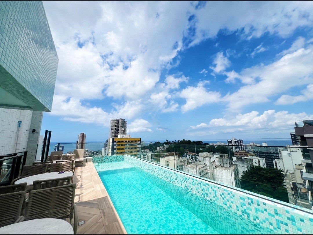 A rooftop area features a shimmering pool with a mosaic tile design. Surrounding the pool, lounge chairs and tables can be seen, offering space for relaxation. The expansive view reveals a cityscape and the distant ocean under a partly cloudy sky.
