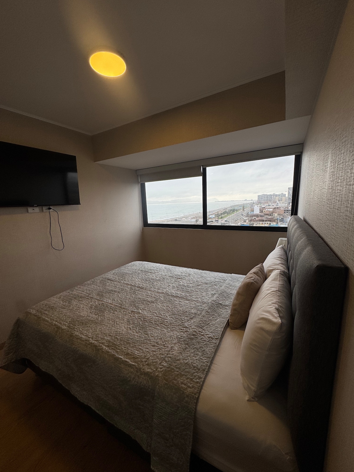 A well-lit bedroom features a spacious bed with neatly arranged linens. Two large windows provide a panoramic view of the sea and surrounding area. A flat-screen TV is mounted on the wall opposite the bed, adding to the room's functionality.