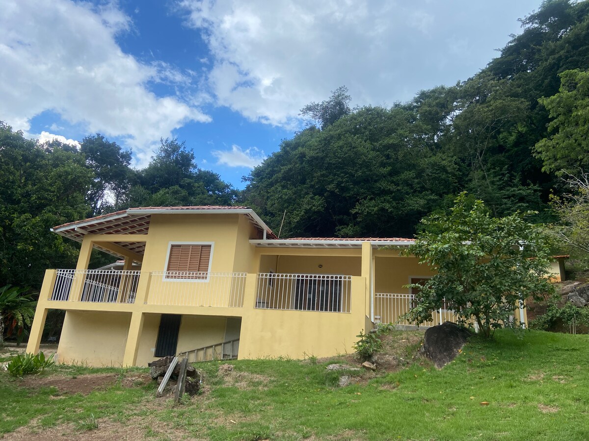 A two-story house is depicted with a yellow exterior complemented by white railings. Surrounded by lush greenery, the structure showcases a spacious covered porch and large windows. The backdrop features a hillside with trees, creating a serene rural atmosphere.
