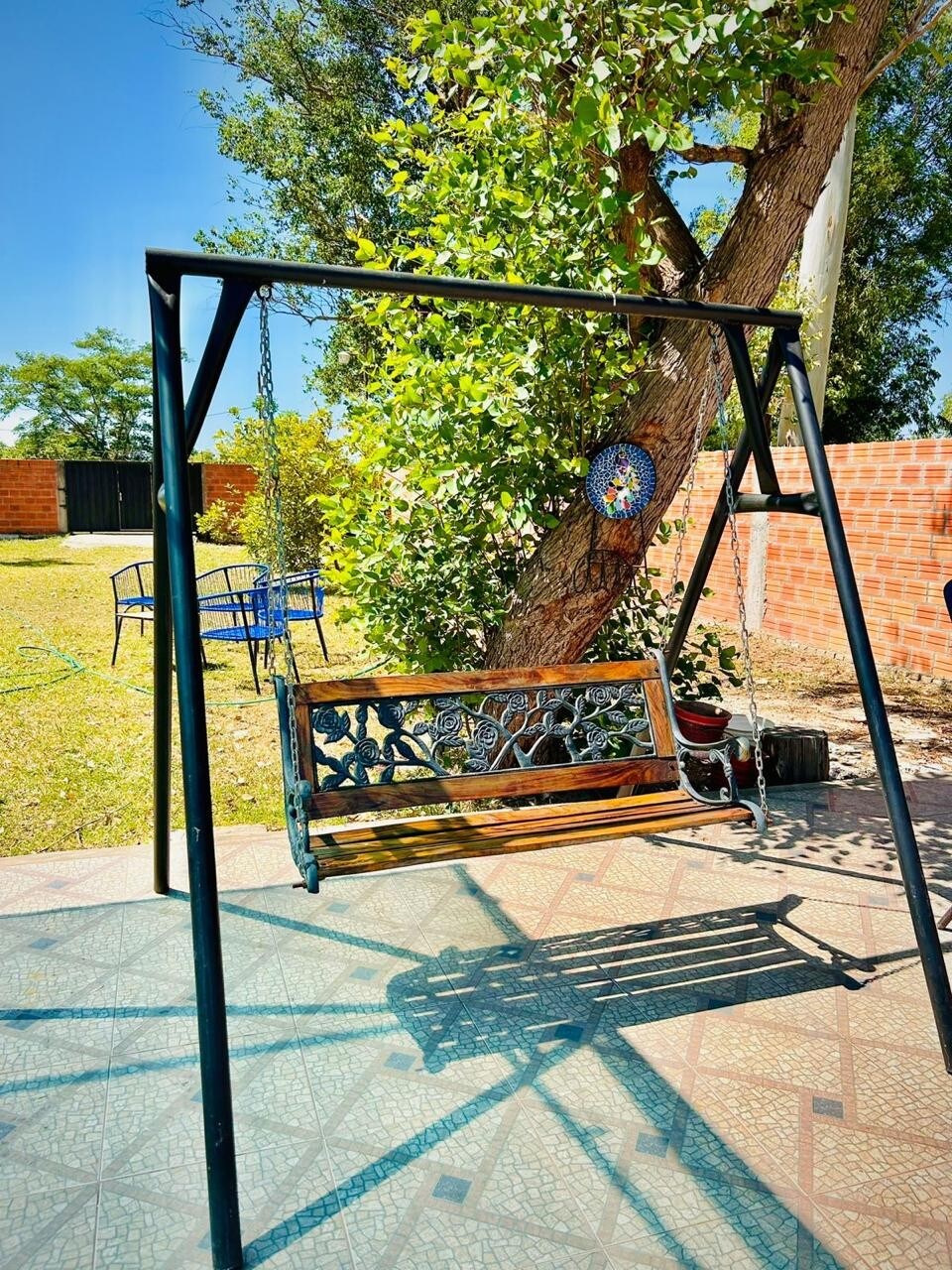 A swing made of wood and metal is suspended under a shady tree, casting a gentle shadow on the patterned patio. Green foliage surrounds the swing, with a colorful decorative piece visible on the tree trunk. Blue chairs can be seen in the background.