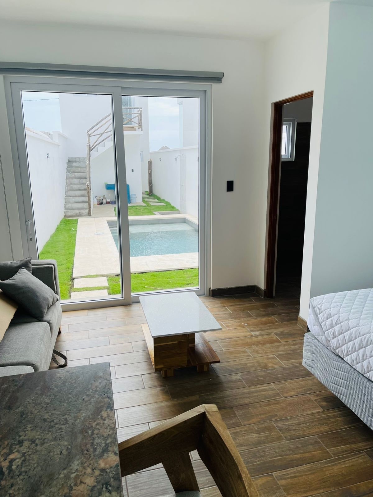 A modern living space features a light grey sofa and a minimalist coffee table. Large glass doors provide natural light and open to a private outdoor area with a pool, surrounded by grass and a staircase leading to additional spaces.