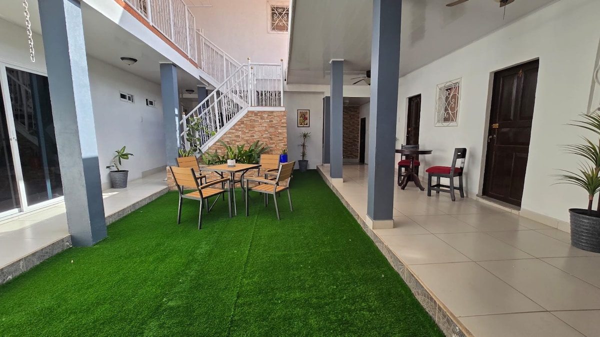 An outdoor area features a manicured lawn bordered by two structures. A set of wooden chairs and a table occupies the green space, inviting relaxation. Nearby, a small potted plant adds a touch of nature, while a doorway leads into the interior of the building.