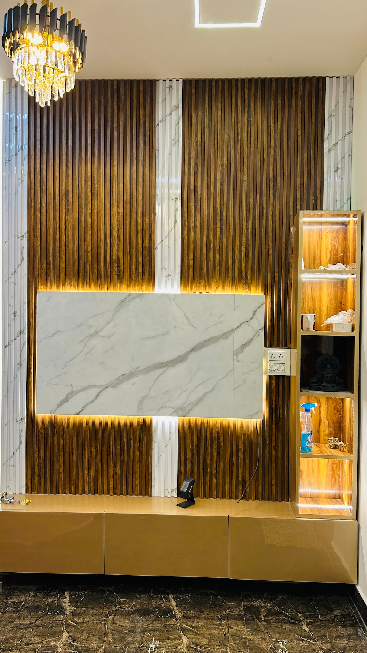 A modern entertainment unit features a large marble wall panel, flanked by wood-paneled accents that create a striking contrast. Soft lighting highlights the textures, while a stylish chandelier hangs above, adding elegance to the space.