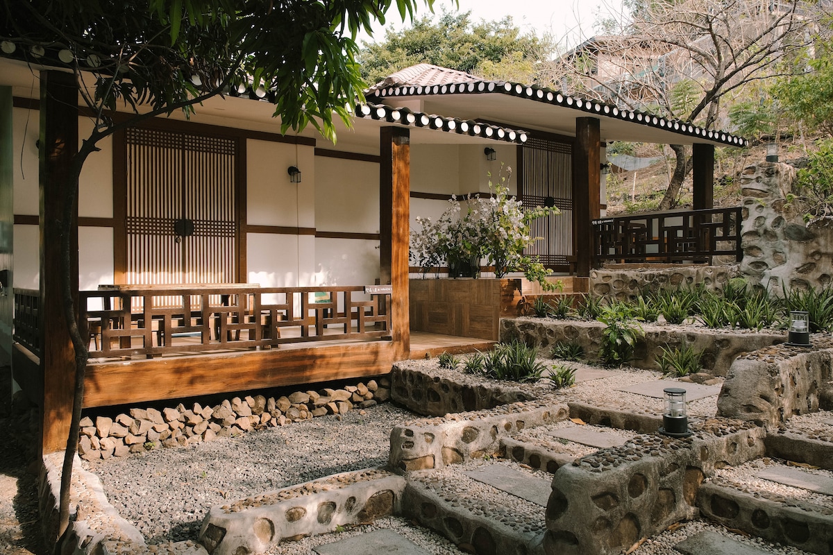 A traditional Korean-style building is framed by neatly arranged stone pathways and landscaped greenery. Wooden pillars support the structure, while large windows offer views of the outdoors. Flowering plants add a natural element to the serene ambiance of the surrounding area.