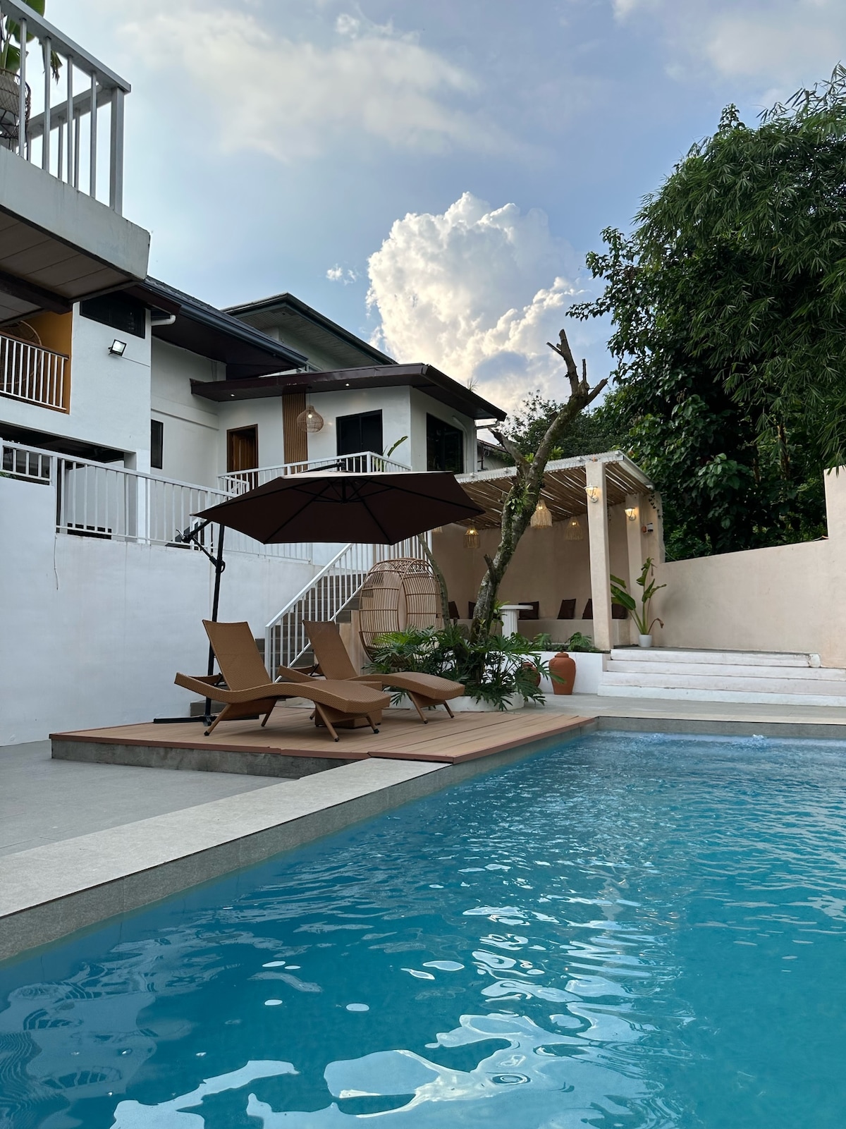An inviting swimming pool is framed by a wooden deck and sun loungers, with an umbrella providing shade. The backdrop features a modern multi-level home and lush greenery, enhancing the tranquil outdoor space. Soft clouds drift in the blue sky above.