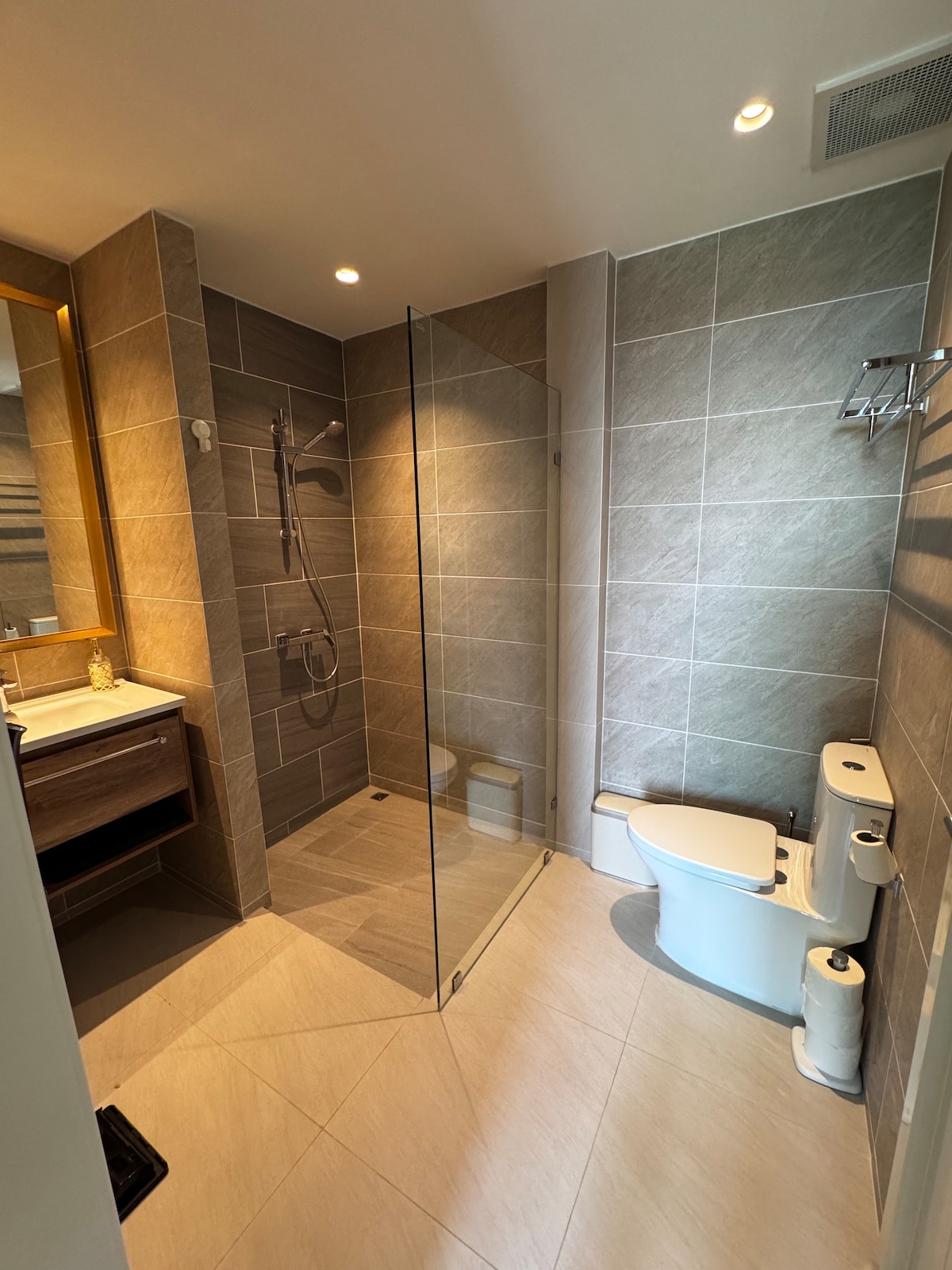 A modern bathroom features a glass-enclosed shower with a rainfall showerhead. Natural light is provided by wall-mounted lights above a vanity with a sink. Neutral-toned tiles create a clean and fresh look, while a toilet is discreetly placed beside the shower area.